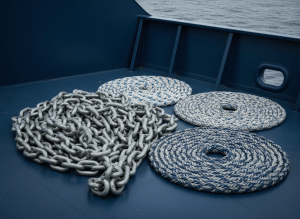 Professional maritime industrial photograph, close-up of heavy galvanized steel anchor chains and coiled marine mooring ropes on a commercial ship deck, deep navy blue and steel gray tones, soft natural daylight, clean minimal composition with negative space, high-end catalog aesthetic, no text, no people